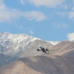 An airplane flies over snow-dusted mountains under a clear blue sky, capturing a sense of adventure and travel.
