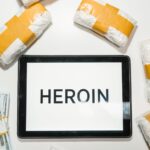 Conceptual image showing packaged heroin, money, and tablet with text in a flat lay arrangement.