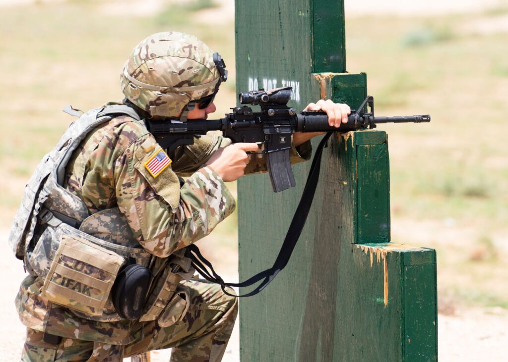 Soldier in camouflage takes aim with a rifle during training, in an outdoor setting.
