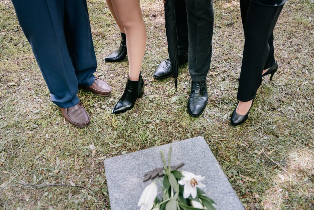 Shoes of mourners gathered around a gravestone with lilies in a cemetery.