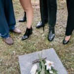 Shoes of mourners gathered around a gravestone with lilies in a cemetery.