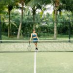A woman poses with a tennis racket on a palm tree-lined tennis court outdoors.