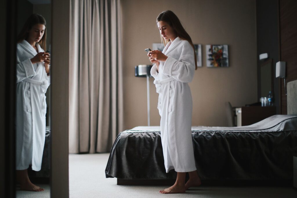Woman in bathrobe looking at smartphone in a modern hotel room, near a mirror.