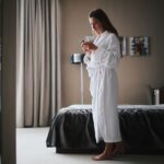 Woman in bathrobe looking at smartphone in a modern hotel room, near a mirror.
