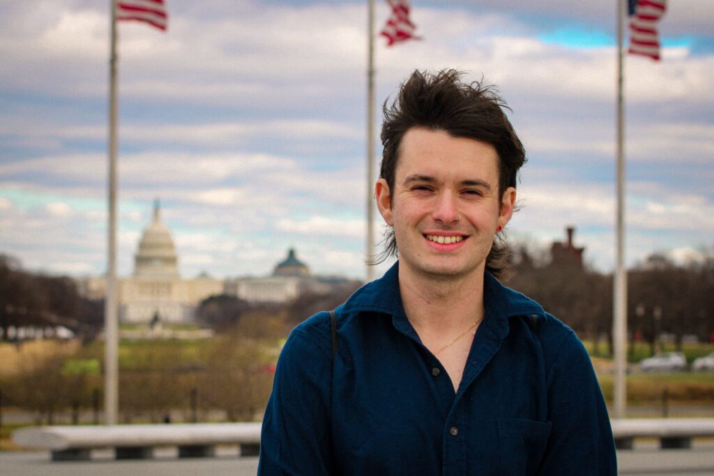 A young man smiling in front of the US Capitol in Washington, DC, with flags in the background.