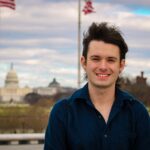 A young man smiling in front of the US Capitol in Washington, DC, with flags in the background.
