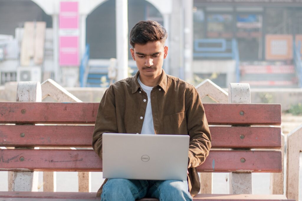 A young man working on his laptop while sitting on an outdoor bench in a city environment.