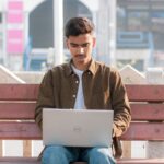 A young man working on his laptop while sitting on an outdoor bench in a city environment.