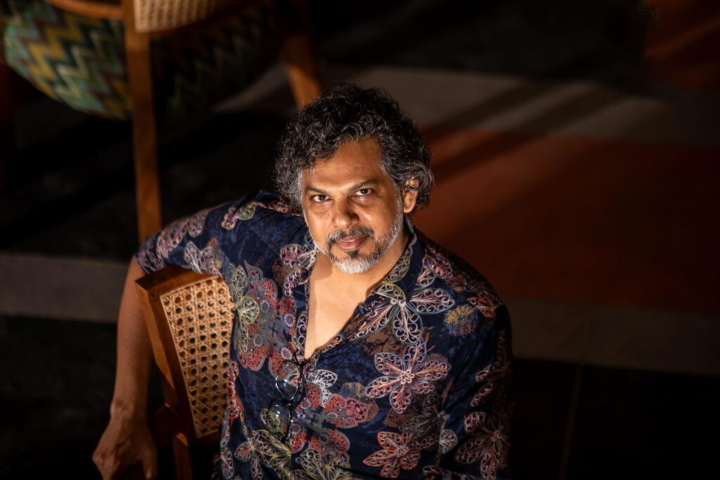 A man with curly hair sits on a wooden chair, wearing a colorful patterned shirt.