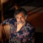 A man with curly hair sits on a wooden chair, wearing a colorful patterned shirt.