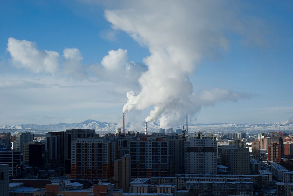 City skyline with industrial smoke set against distant mountains under a clear sky.