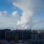 City skyline with industrial smoke set against distant mountains under a clear sky.