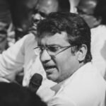 Black and white image of a man with eyeglasses speaking into a microphone at an event.