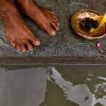 Barefoot ritual beside the water featuring a lit offering on a brass plate.