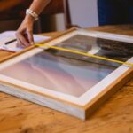 Hands using a tape measure to size a wooden picture frame on a wooden table indoors.
