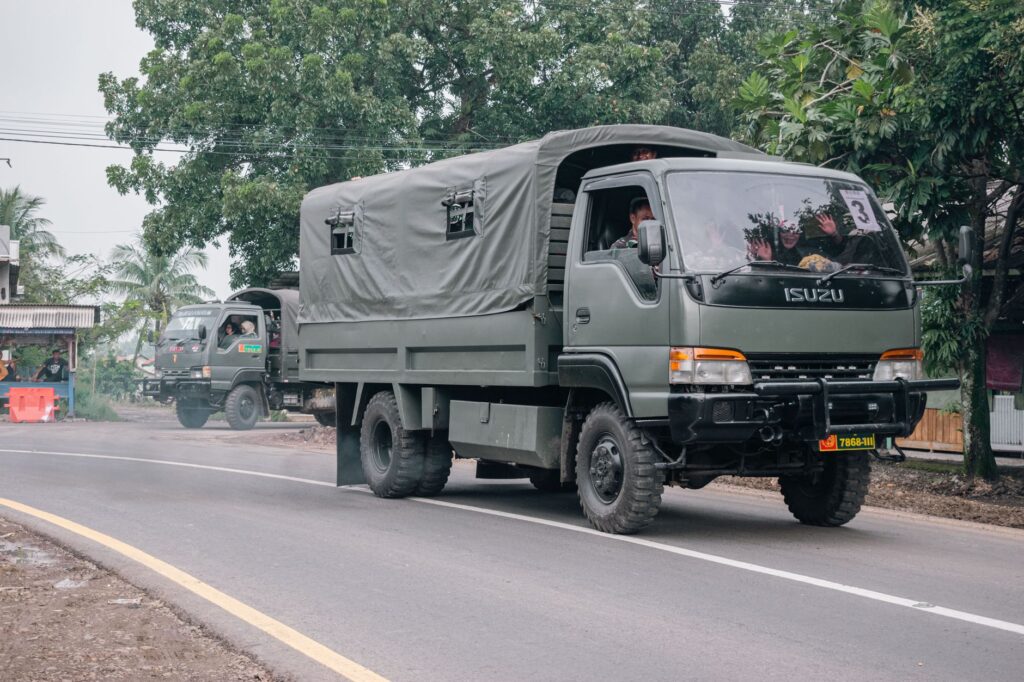 Military trucks traveling on a rural road surrounded by trees and local buildings.
