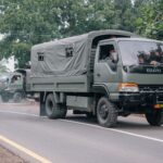 Military trucks traveling on a rural road surrounded by trees and local buildings.