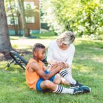 A woman comforts an injured child beside a bicycle in a sunny park setting.
