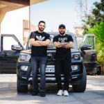 Two men with crossed arms standing in front of an SUV in urban Jaipur, Rajasthan.