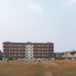 Cricket match in progress at a sports field in Delhi, India, with players and spectators under clear skies.