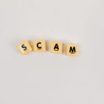 Four Scrabble tiles forming the word 'SCAM' on a neutral background, highlighting concepts like deceit.