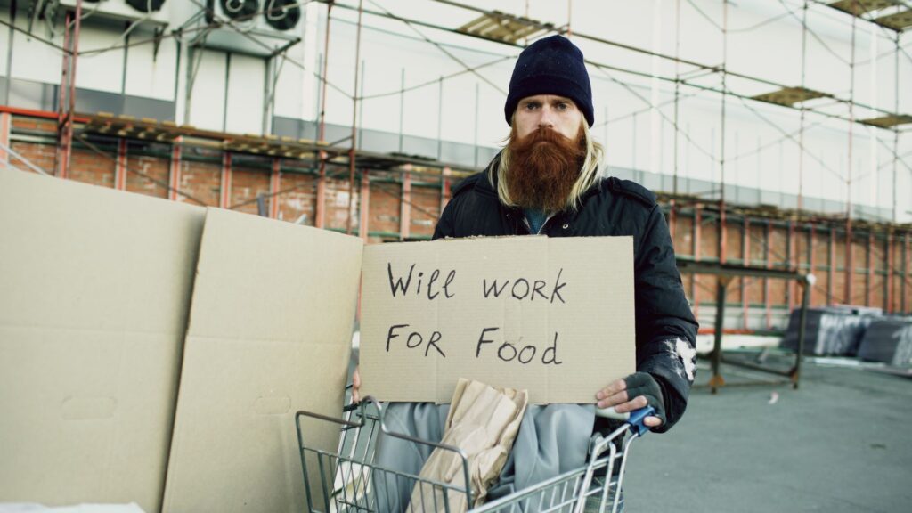 A homeless man with a sign 'Will work for food' in an urban setting.