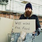 A homeless man with a sign 'Will work for food' in an urban setting.