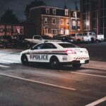 A police car drives through an urban street at night illuminated by streetlights.