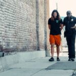Police officer walking with handcuffed man along urban street with brick wall.