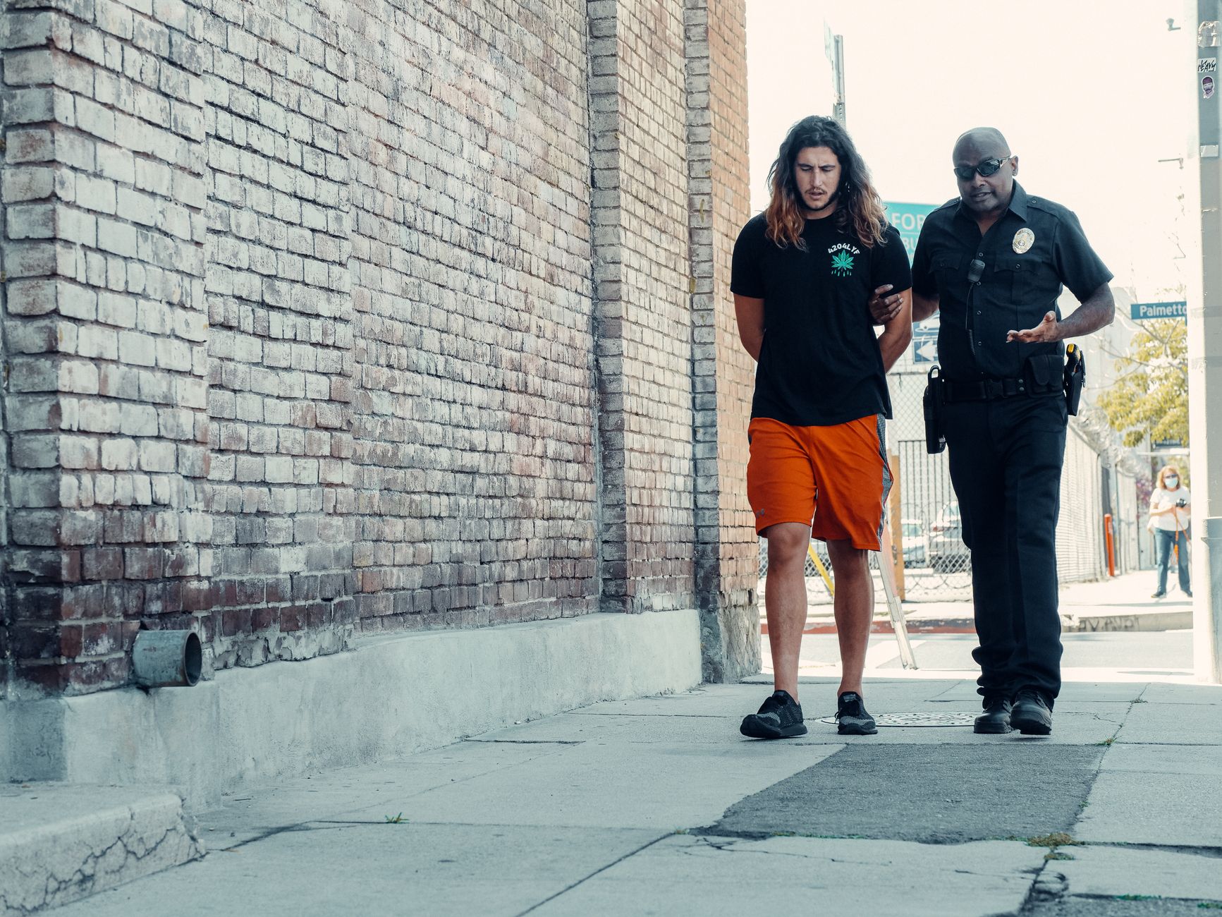 Police officer walking with handcuffed man along urban street with brick wall.