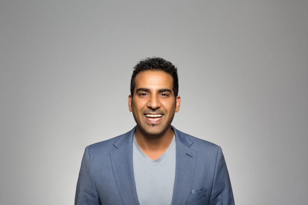 Portrait of a joyful man in a blue blazer, smiling against a plain gray backdrop.