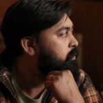 Profile portrait of a bearded South Asian man deep in thought, wearing casual plaid indoors.