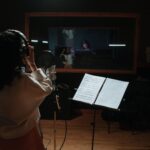 A female singer records vocals in a professional music studio with a microphone and sheet music.