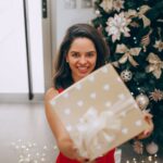 Smiling woman holding a Christmas gift in front of a decorated tree indoors.
