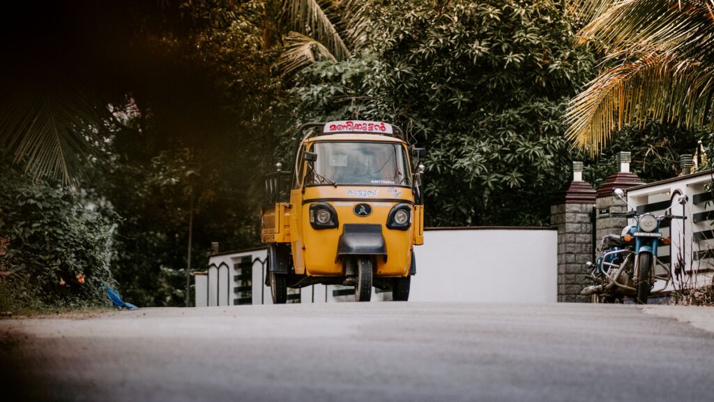 A yellow auto rickshaw on a scenic street in Kerala, surrounded by lush greenery and coconut palms.
