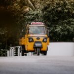 A yellow auto rickshaw on a scenic street in Kerala, surrounded by lush greenery and coconut palms.