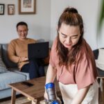 A woman smiling while vacuuming a living room, with a man working on a laptop in the background.