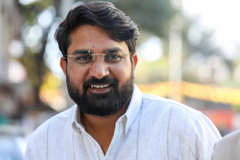 Smiling man with beard and glasses in an outdoor setting, exuding warmth and friendliness.