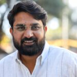 Smiling man with beard and glasses in an outdoor setting, exuding warmth and friendliness.