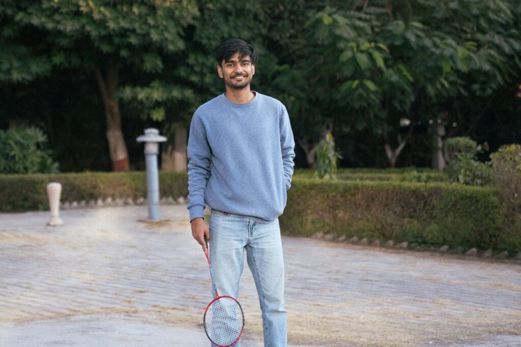 Smiling young man with a badminton racket in a park setting, enjoying leisure time.