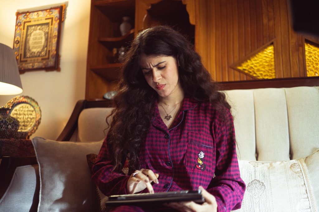 A woman in red plaid pajamas focuses on her tablet while relaxing on a sofa indoors.
