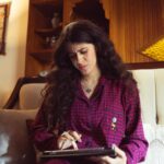 A woman in red plaid pajamas focuses on her tablet while relaxing on a sofa indoors.