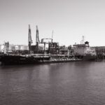 Monochrome photo of a cargo ship docked at an industrial port with cranes in the background.