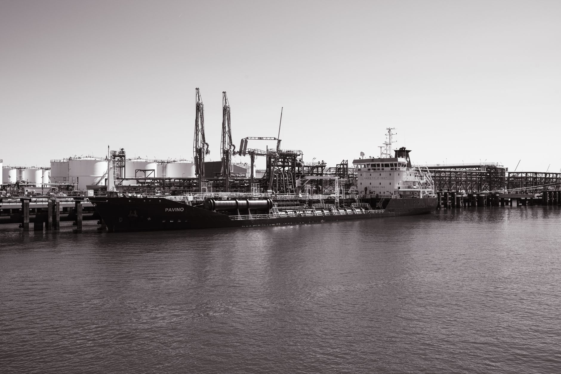 Monochrome photo of a cargo ship docked at an industrial port with cranes in the background.