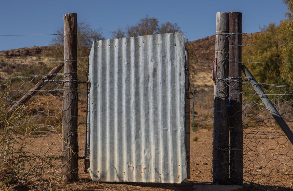 Corrugated metal gate framed by wooden posts in a dry, rugged landscape under clear skies.