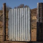 Corrugated metal gate framed by wooden posts in a dry, rugged landscape under clear skies.