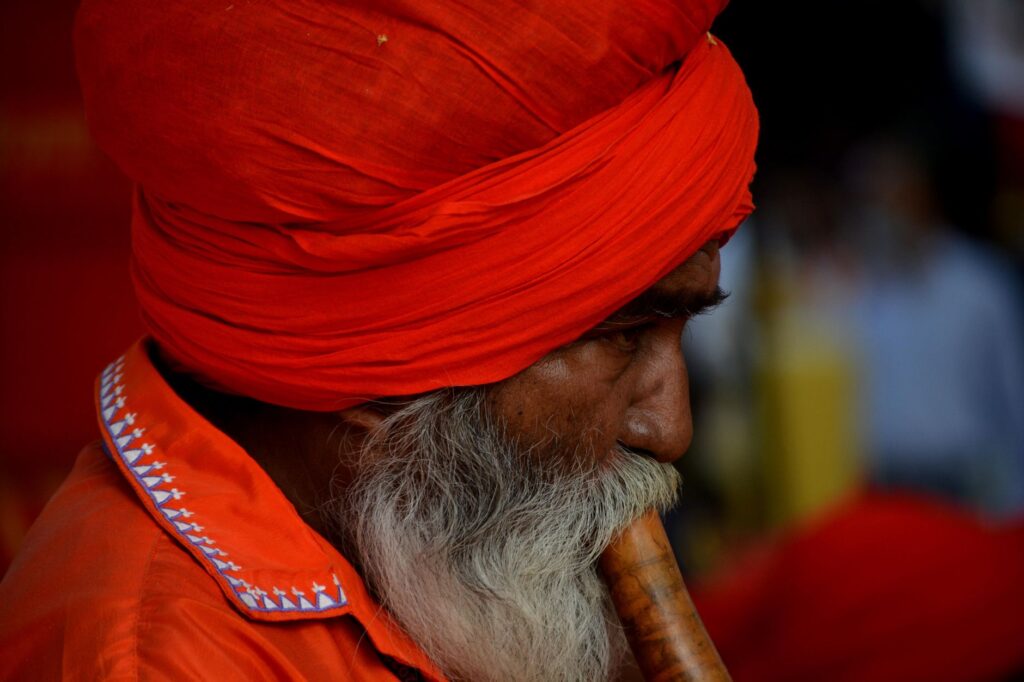 Elderly Indian man in vibrant orange attire playing a traditional flute at a cultural fair.