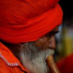 Elderly Indian man in vibrant orange attire playing a traditional flute at a cultural fair.