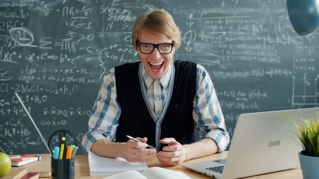 Smiling male teacher in a classroom with a mathematical blackboard and laptop.