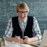 Smiling male teacher in a classroom with a mathematical blackboard and laptop.
